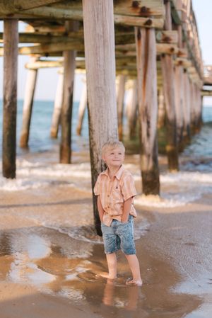 A young child wades in shallow water under a wooden pier at the beach on a sunny day.