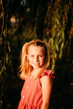 Young person in coral dress poses under willow branches at golden hour.