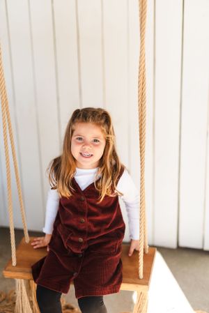 A child in a burgundy corduroy dress sits happily on a wooden rope swing.
