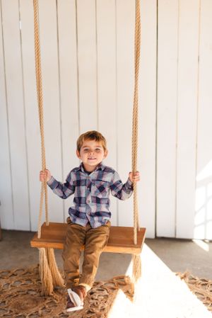 A young child in plaid shirt and khakis smiles while playing on a wooden swing.