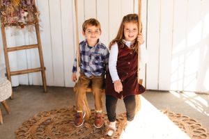 Two children sit together on a wooden rope swing against a white wall.