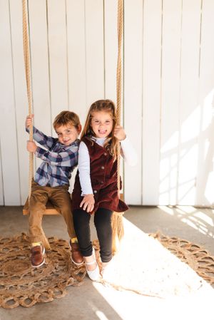 Two playful children share a wooden swing in front of a white wall with natural lighting.