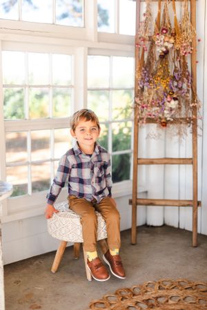 Child sitting on white stool in bright sunlit room decorated with dried flowers and wooden ladder.