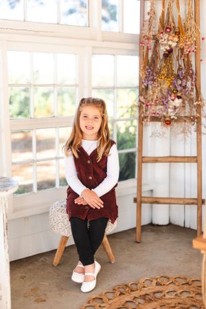 Child sitting on white stool wearing burgundy vest and white shirt in sunny corner room.