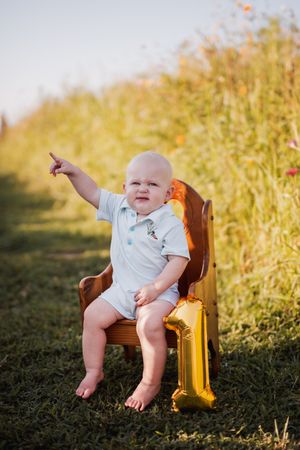 A happy baby sits in a wooden chair outdoors surrounded by golden sunlit grass pointing into the distance.