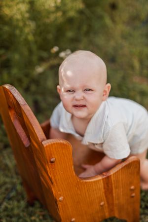 A baby in white clothing peeks over the edge of a wooden chair in an outdoor setting.