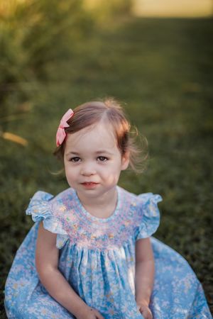 A child in a light blue floral smock dress with ruffled sleeves sits on grass during golden hour.