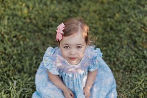 A child in a light blue dress with cape and crown sits in green grass looking at the camera playfully.