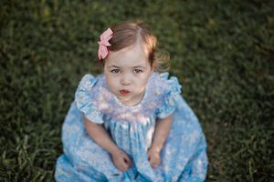 Toddler in a blue dress with ruffles and a pink bow poses on grass during an outdoor photo session.