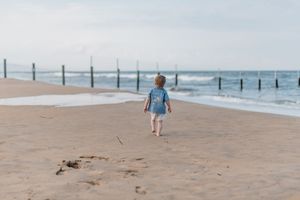 Person in blue shirt and backpack walks along vast empty beach with wooden posts on cloudy day.