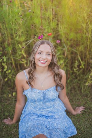 A smiling figure in a flowing blue and white floral dress poses against a natural green background outdoors.