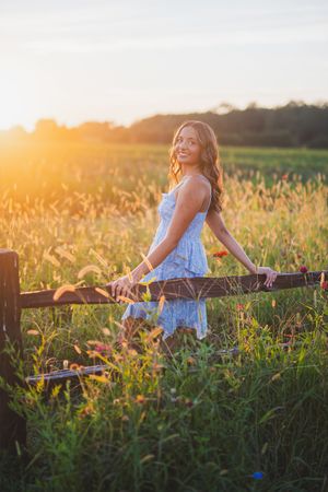 A figure sits on a rustic wooden fence surrounded by wildflowers at golden hour in a pastoral countryside setting.