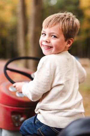 Child in cream sweater sitting on red tractor in autumn setting.