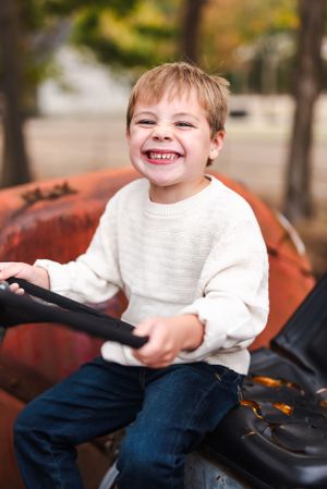 Child in white sweater smiling while playing on red tractor outdoors.