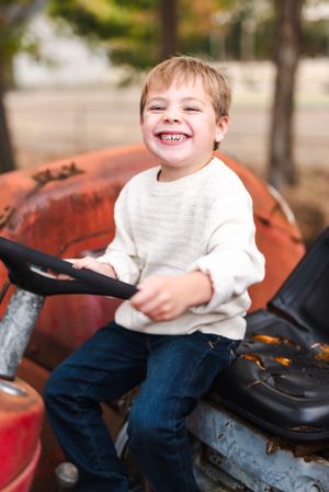 A child with a big smile sits on a vintage red tractor wearing a white sweater and jeans.