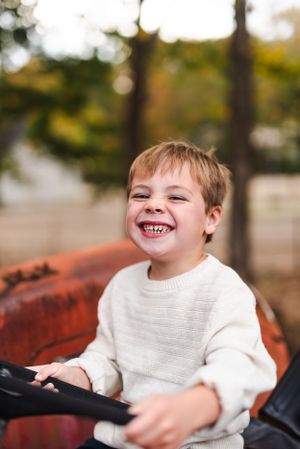 Happy child playing on red tractor with autumn trees in background.