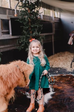 A child in a green dress sits with a large golden-colored dog on a wooden bench in an indoor setting.