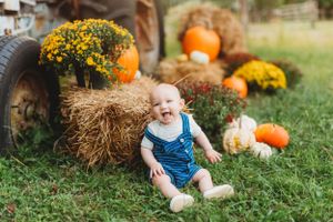Baby in overalls sits among autumn decorations with pumpkins, hay bales, and yellow mums.