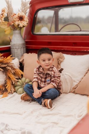 Cozy fall photo setup in vintage red truck bed with plaid shirt, pillows, pumpkins and autumn floral arrangements.