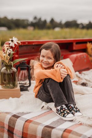 A cozy fall picnic setup with orange sweater, plaid blanket, mason jar flowers and lantern in red truck bed.