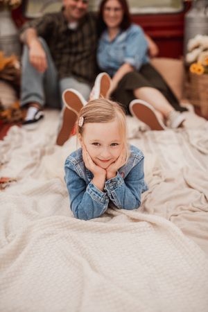Child wearing jean jacket lies on white bedding during a casual indoor photo shoot.