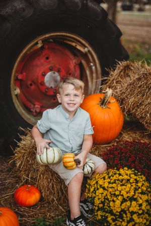 Fall scene with child sitting on hay bale next to large pumpkins and tractor wheel.