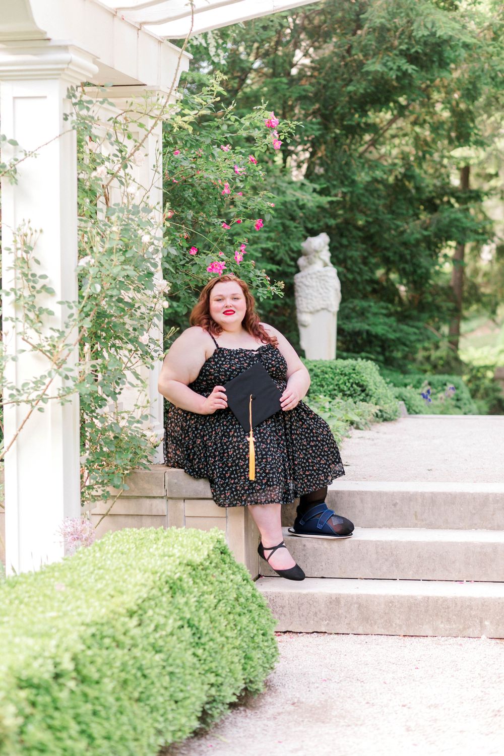 Woman in black dress holding graduation cap on garden steps with roses blooming in background.