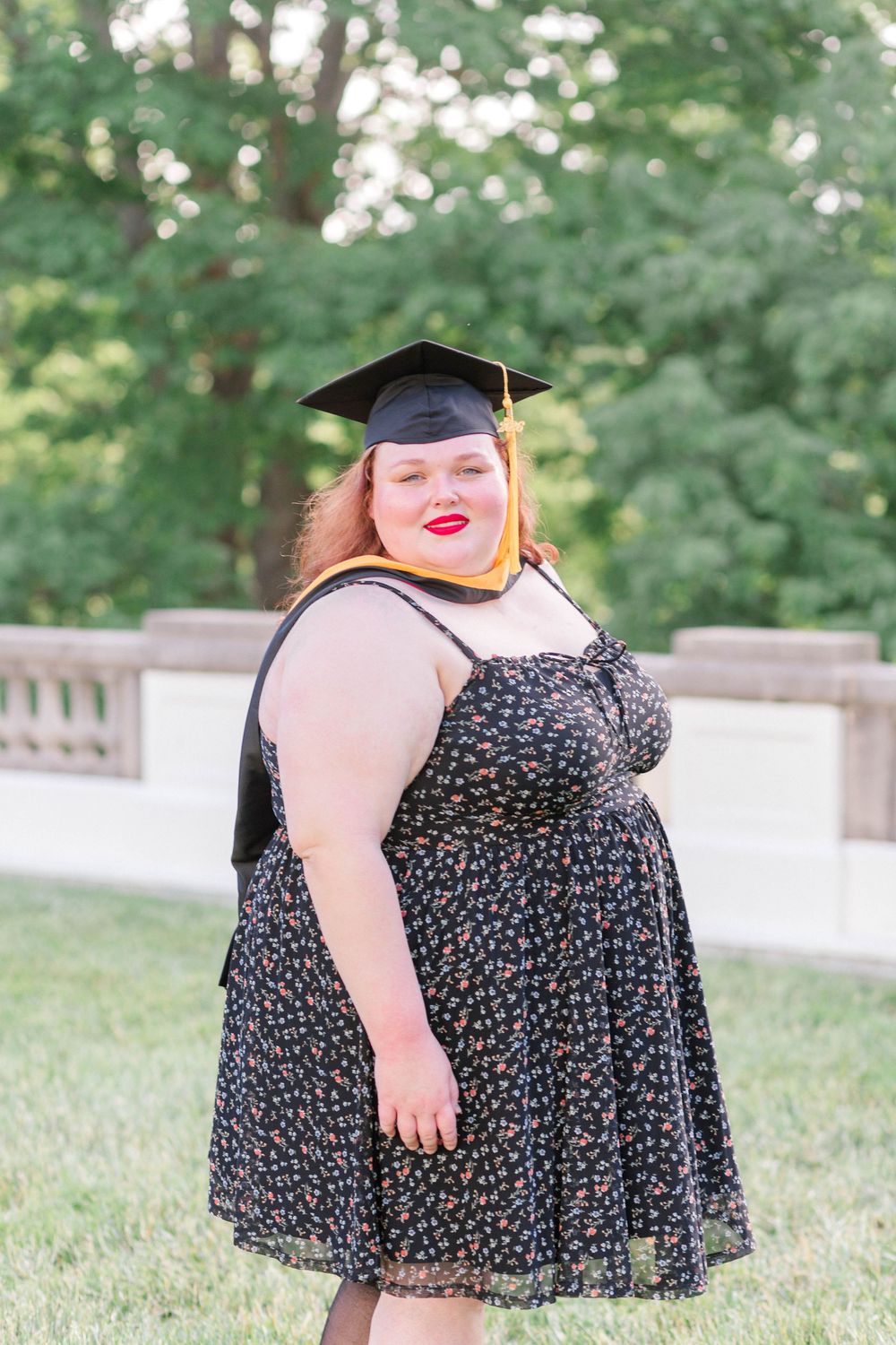 A plus-size woman in a floral dress and graduation cap smiles outdoors, celebrating her academic achievement.