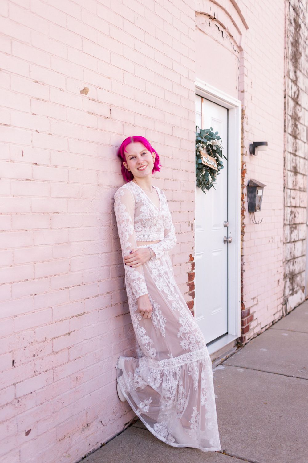 Woman with pink hair in white lace maxi dress leaning against pink brick wall outdoors.