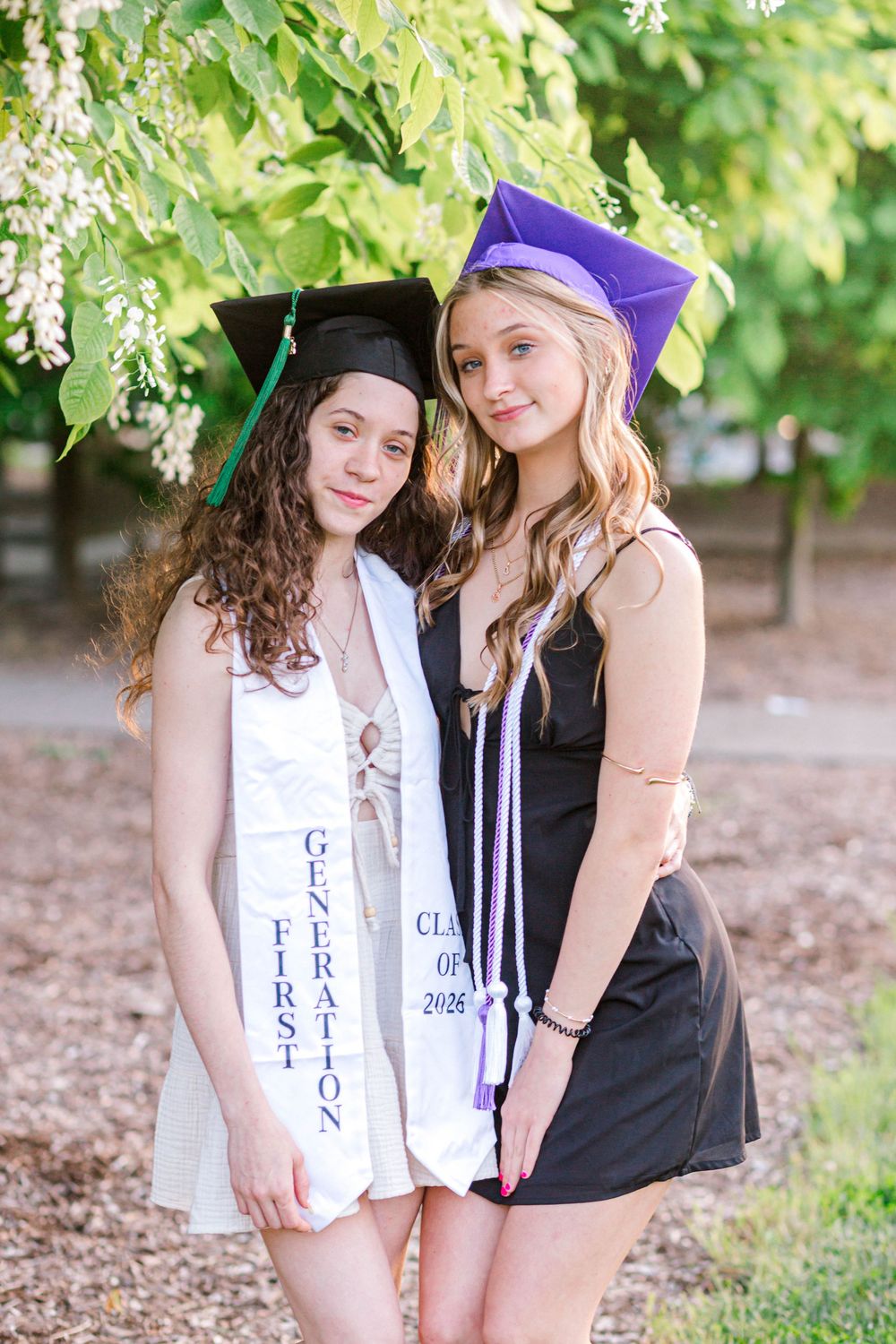 Two young women in graduation caps and stoles, one black and one purple, posing together outdoors under green trees.