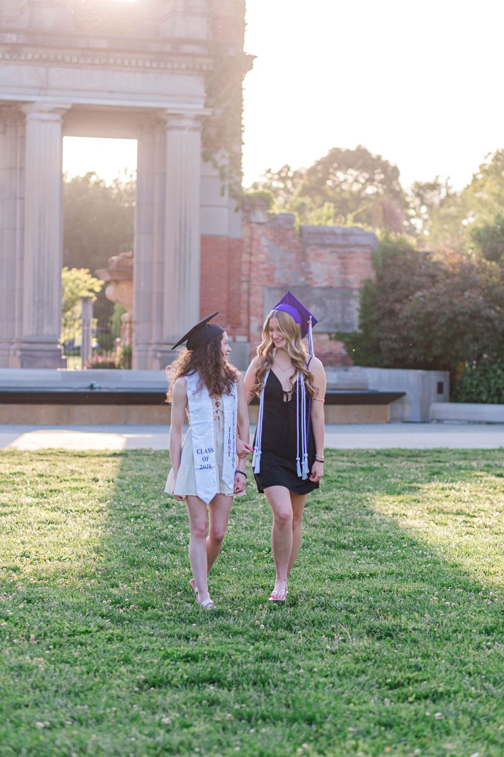 Two female graduates in caps and gowns walk on green grass near classical columns during golden hour.
