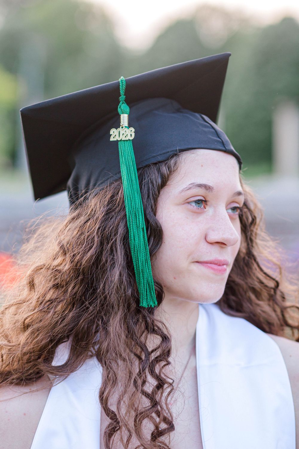Young woman wearing black graduation cap with green 2026 tassel and white gown, smiling outdoors at graduation ceremony.