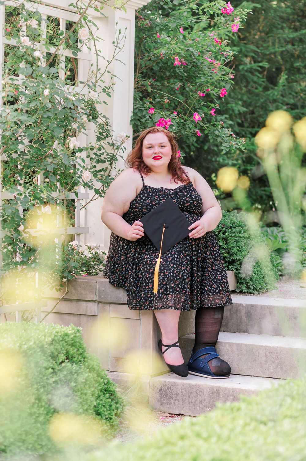 Graduate in floral dress holding mortarboard cap on garden steps surrounded by lush greenery and pink roses.