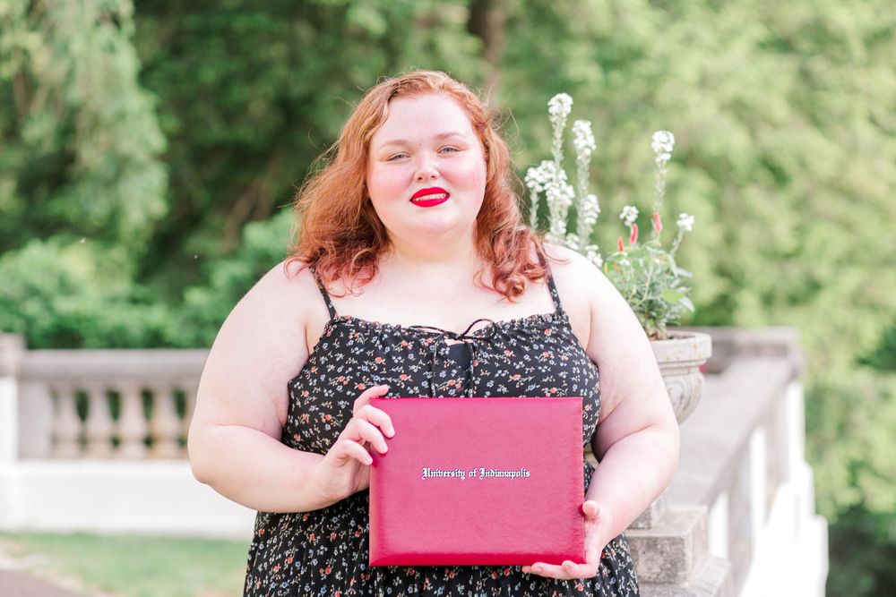 Young woman with red hair holding a pink University diploma folder, wearing a floral dress outdoors.
