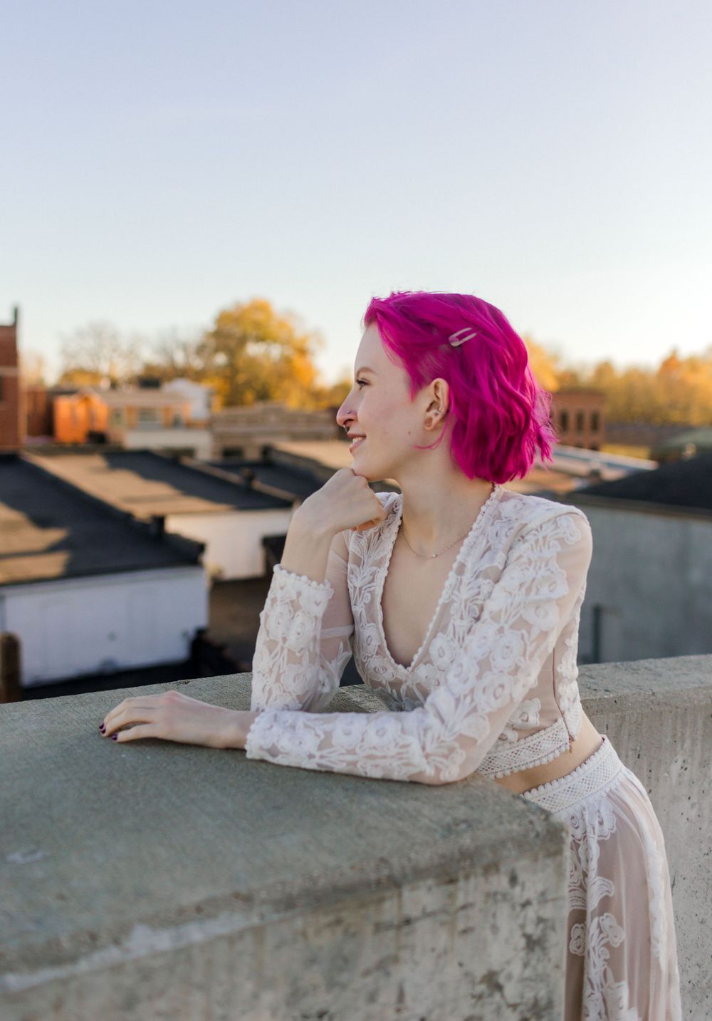 Woman with vibrant pink hair in white lace outfit leaning on rooftop ledge, smiling at golden hour sunset.