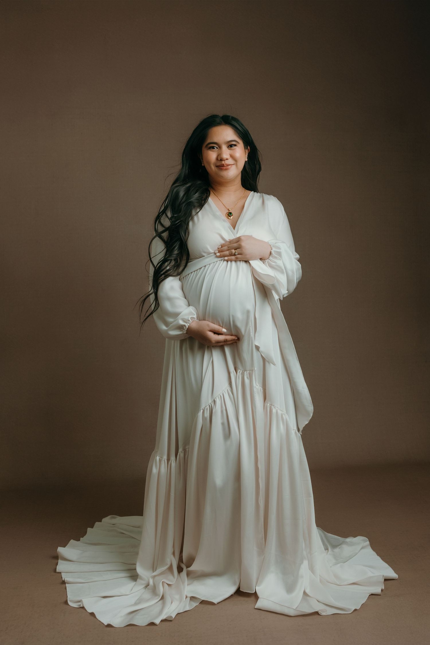 Pregnant woman in elegant white flowing maternity gown cradling baby bump in professional studio portrait in Victoria BC.