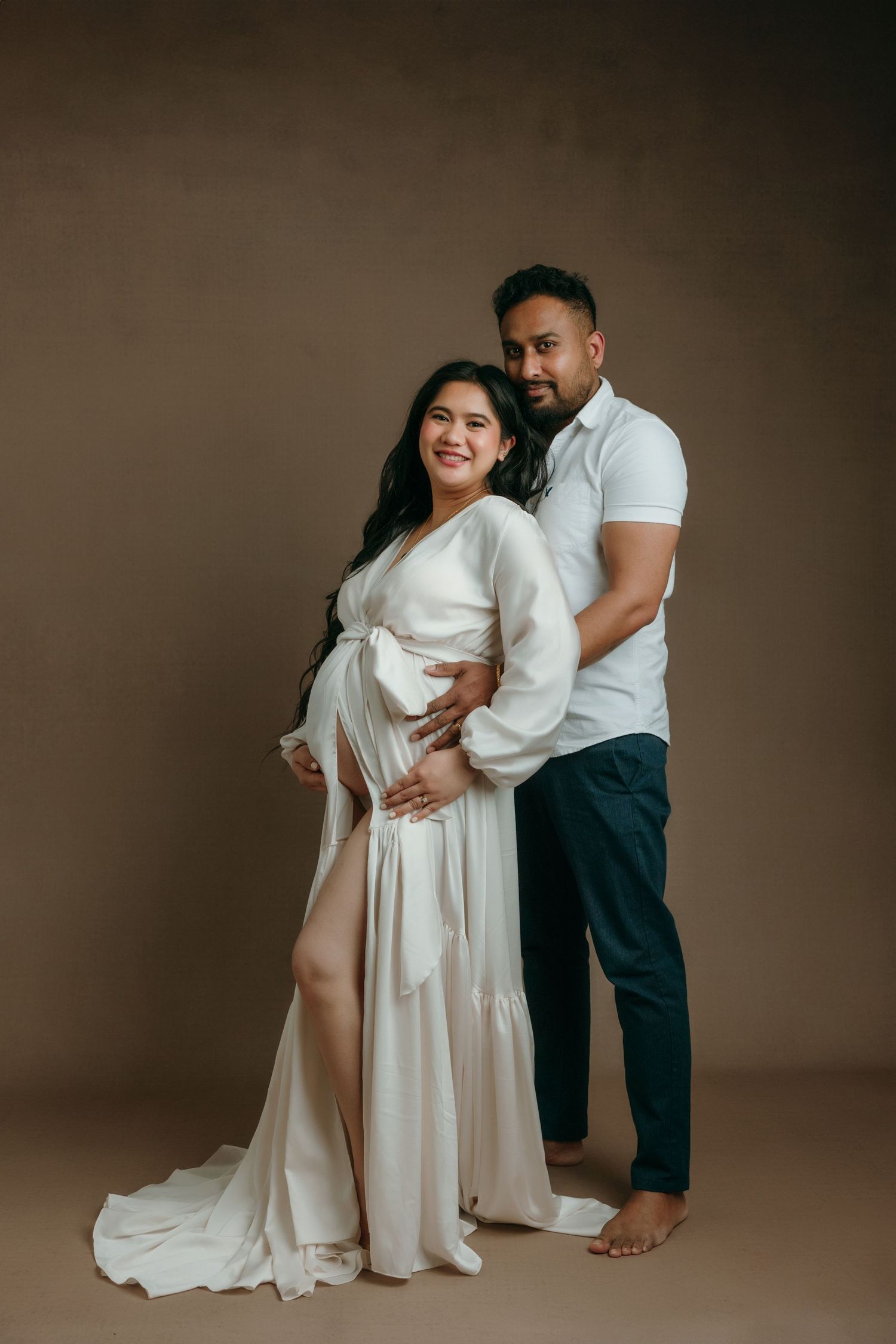 Couple maternity photo, pregnant woman in white gown embraced by partner in maternity studio in Victoria BC.