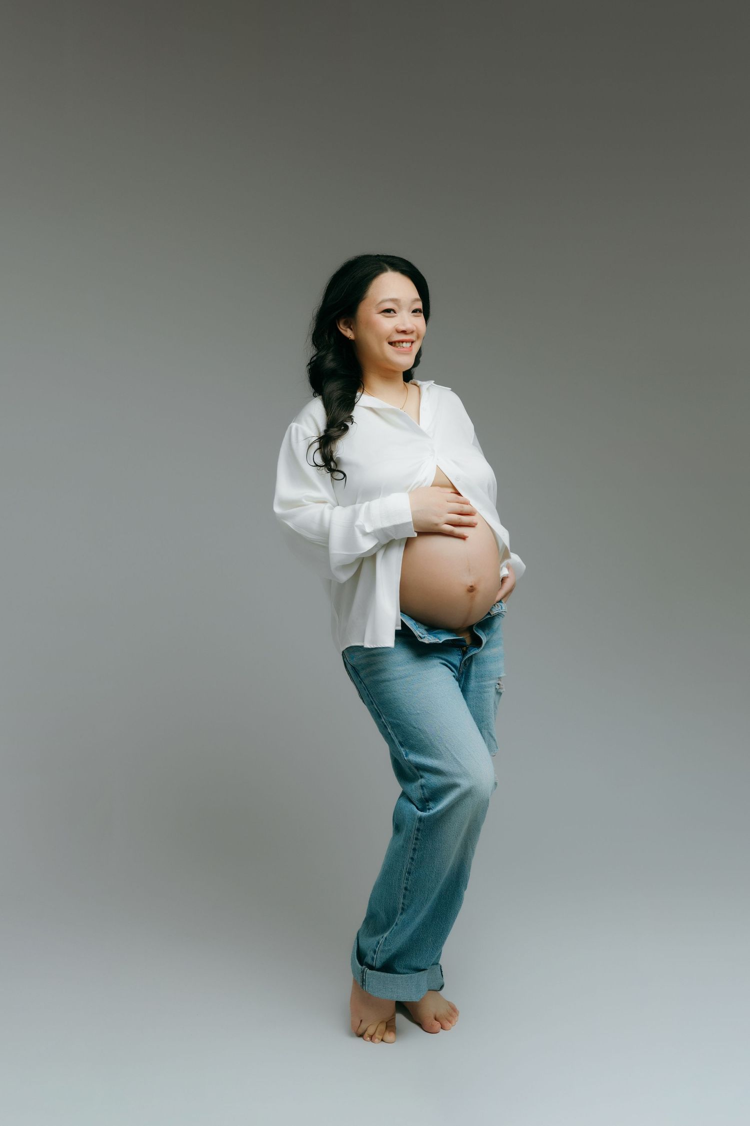 Smiling pregnant woman in white shirt and jeans holding baby bump, barefoot at the maternity studio in Victoria BC