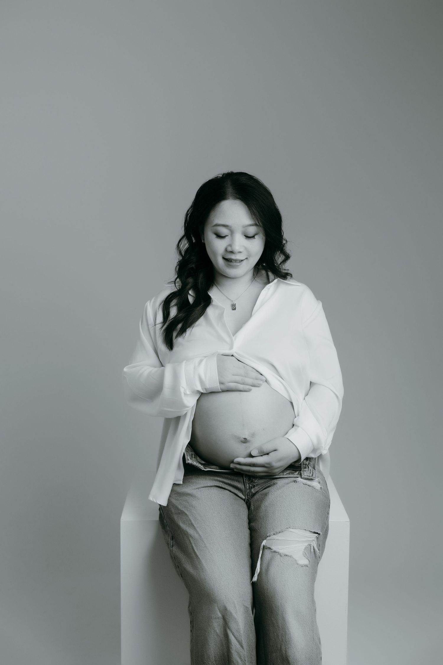 Black and white photo of a pregnant woman in white shirt and jeans cradling her baby bump at the maternity studio in Victoria BC