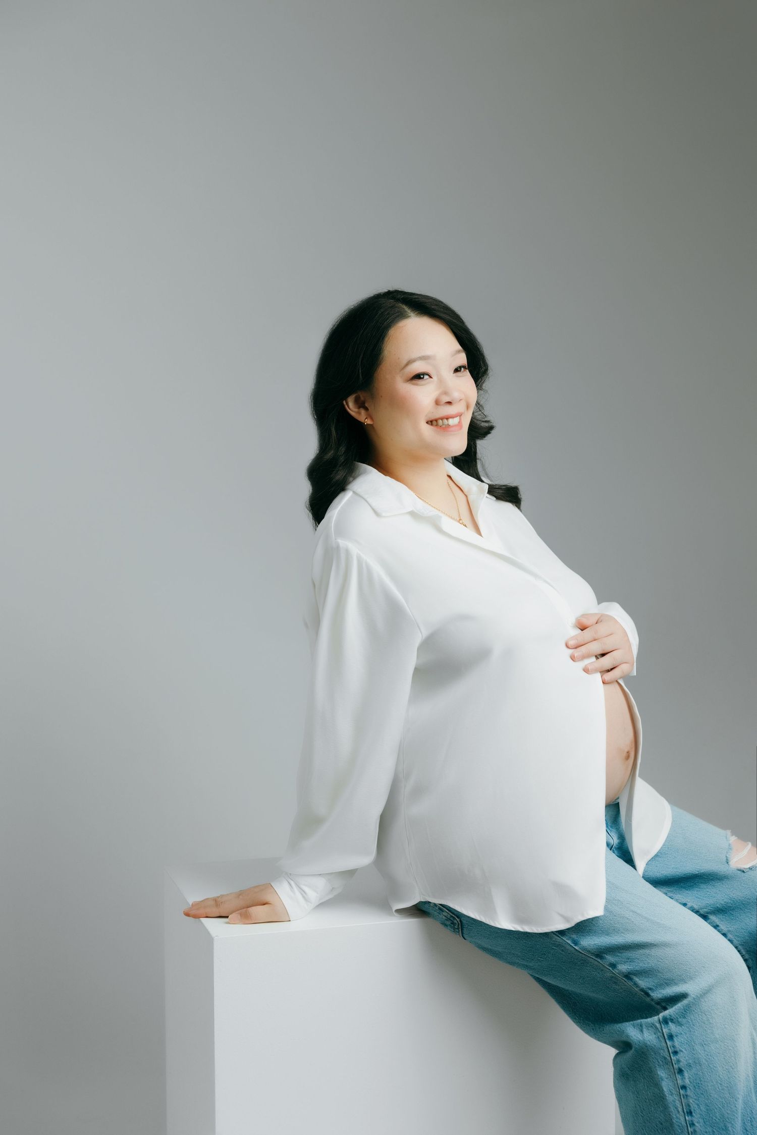 Smiling pregnant woman in white shirt and jeans sitting on white cube, cradling baby bump in maternity studio in Victoria BC.