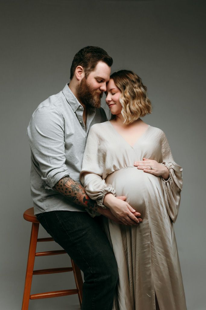 Intimate maternity photo featuring couple in tender moment against grey backdrop with wooden stool at the studio in victoria bc.