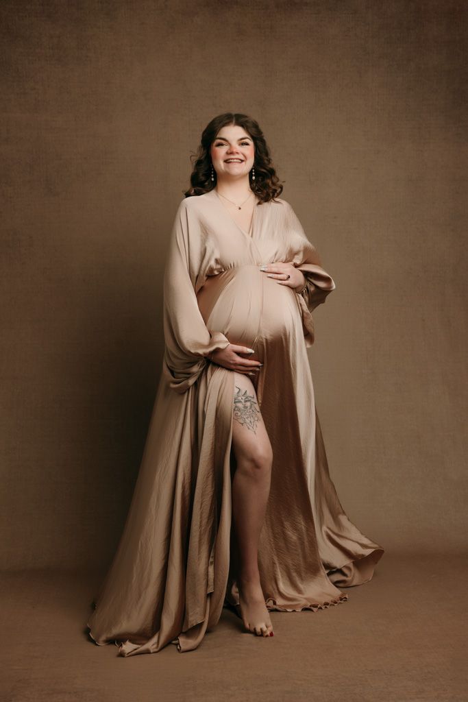 An elegant maternity portrait features a woman in a flowing beige dress posing against a brown backdrop at studio in victoria bc