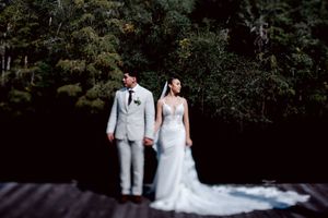 A couple celebrates in formal wedding attire under a canopy of lush green forest trees and dappled sunlight.