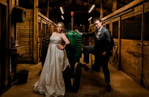 Inside a rustic wooden barn aisle, a couple in wedding attire shares a playful moment with a tractor.