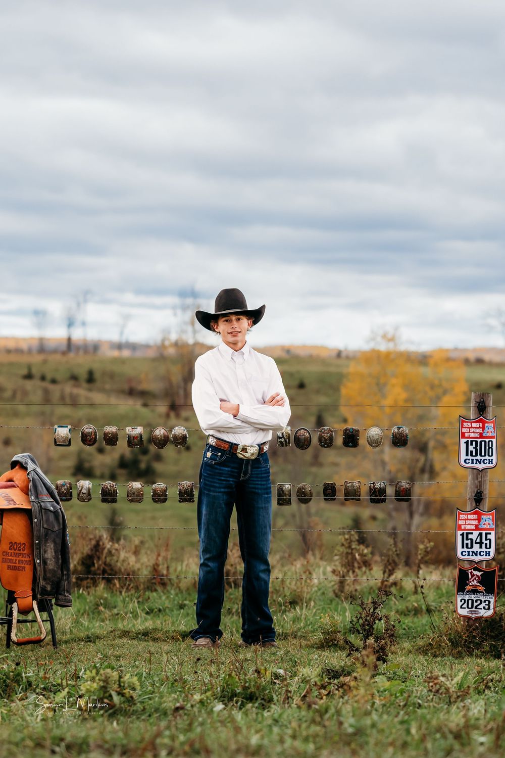 young cowboy posing with championship belt buckles on fence in field with saddles on side