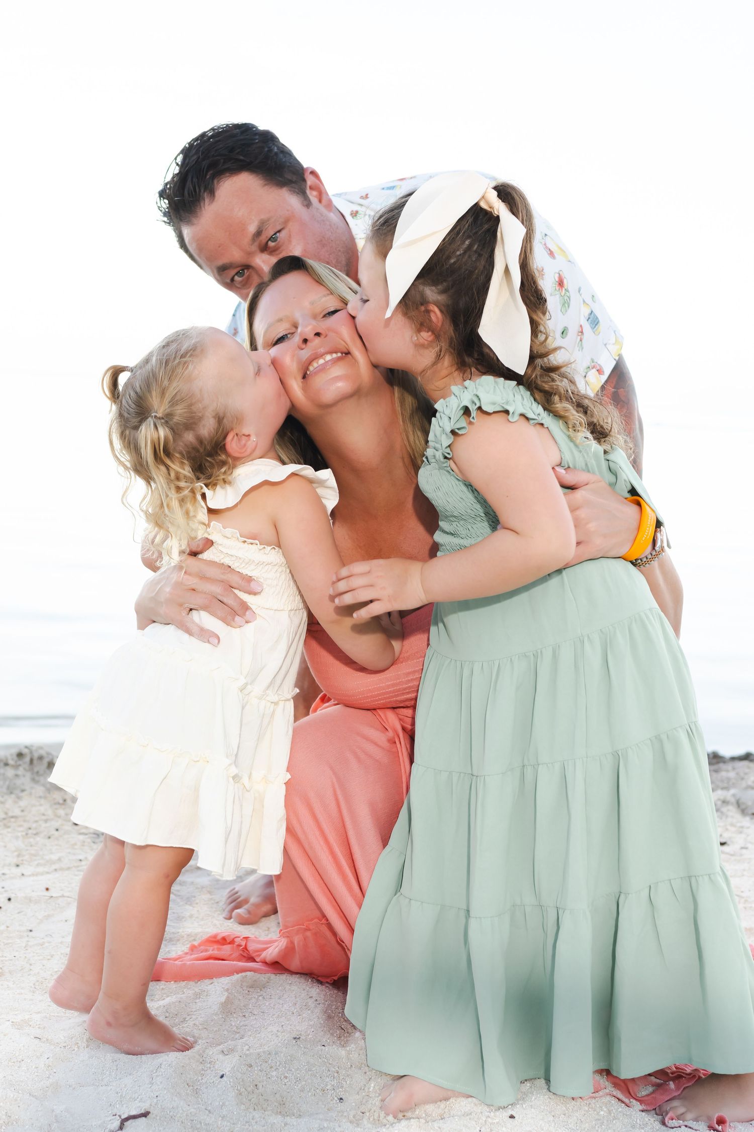 A sweet family moment captured on the beach as they share kisses and hugs while wearing pastel summer dresses.