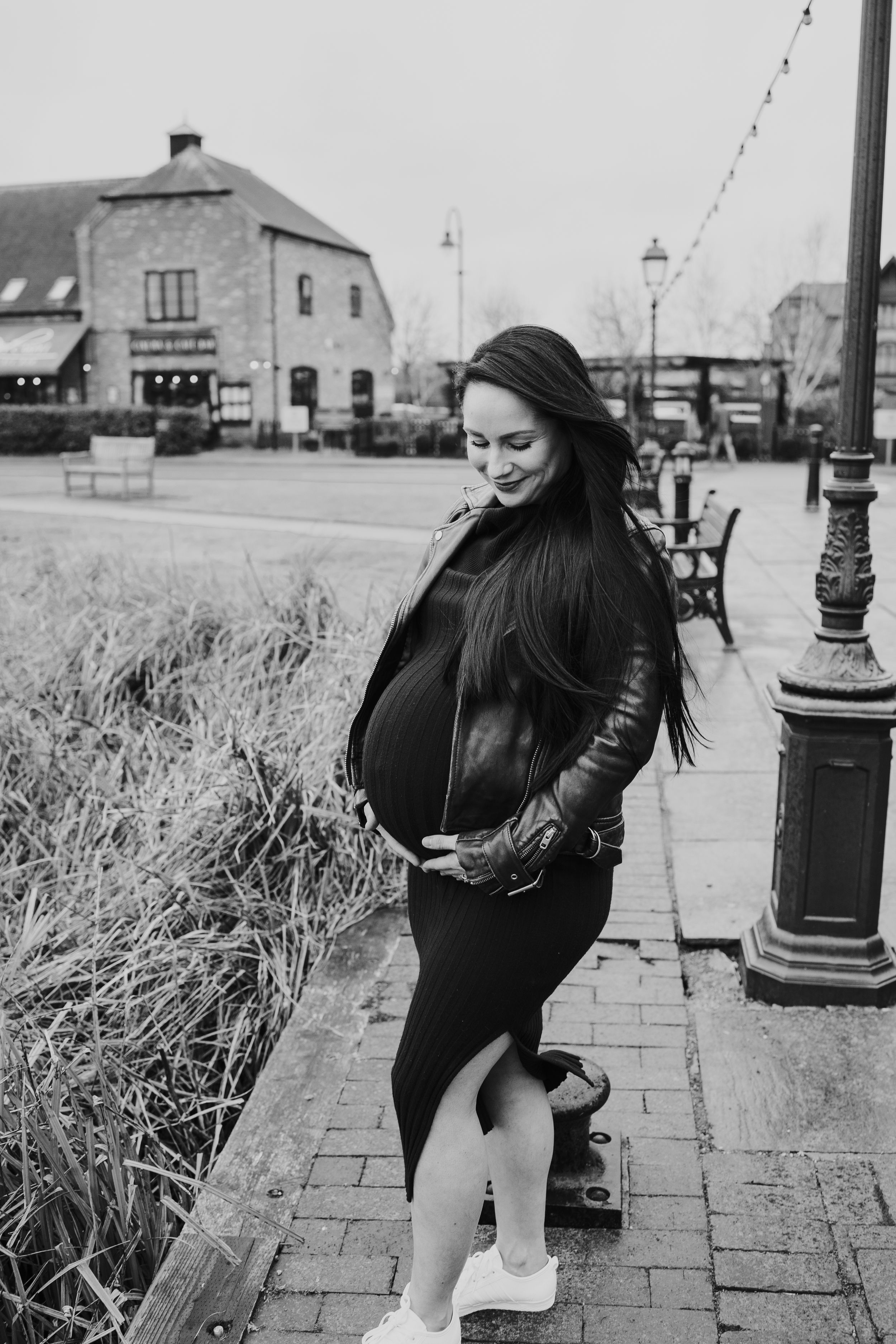 A pregnant woman in a leather jacket stands on a brick sidewalk next to a vintage lamppost in a black and white photo.