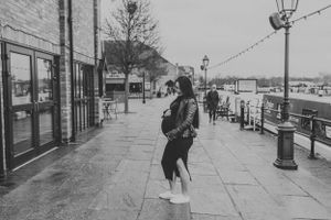 Black and white photograph showing a person standing on a waterfront promenade with storefronts and string lights.