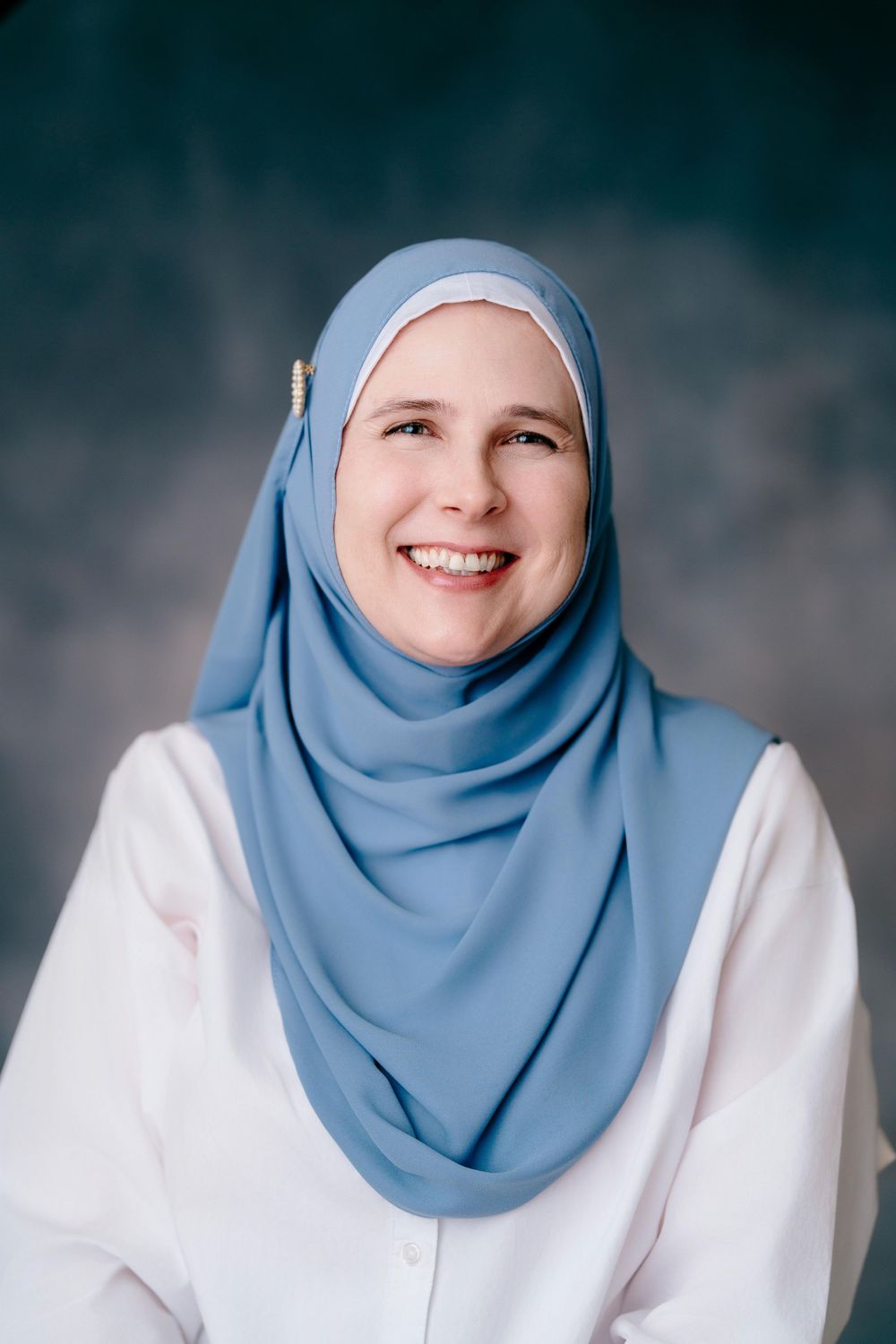 Smiling woman wearing a light blue hijab and white shirt poses for a portrait against a dark background.