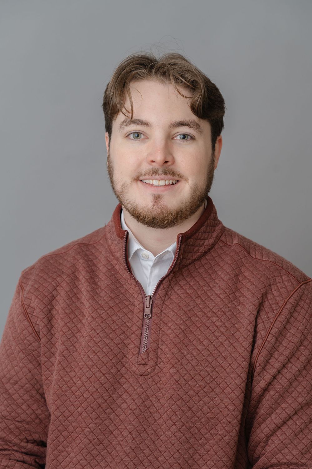 Young man with beard wearing burgundy quilted quarter-zip pullover poses for casual professional headshot.